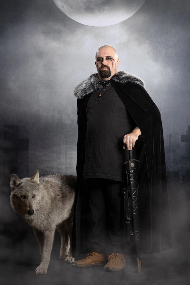 A stern man in black cloak with sword, standing beside a wolf in mist.
